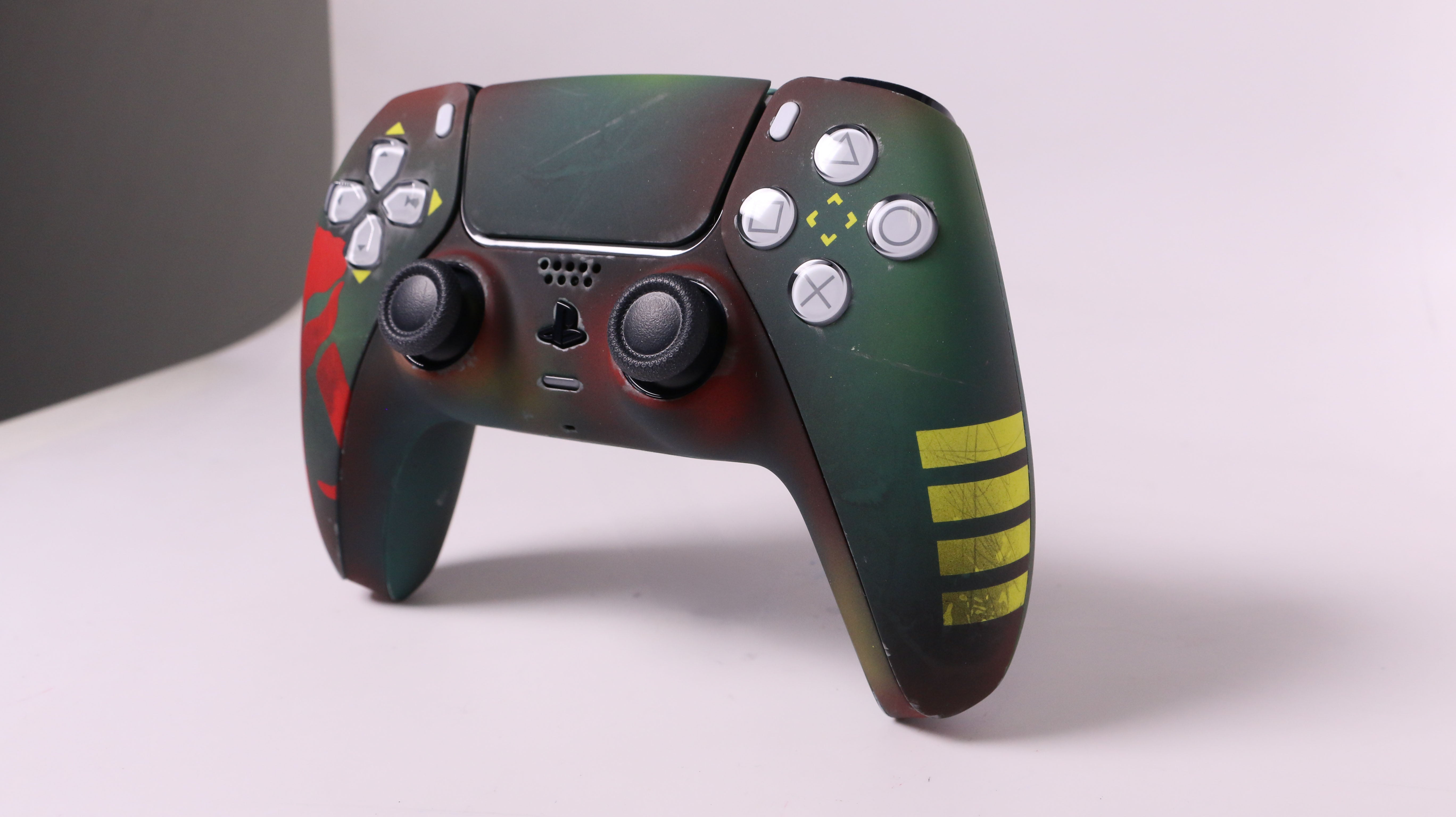 A custom-decal PlayStation 5 controller with a dark green and black camouflage pattern, red splatters, yellow stripes on the right grip, and a clear stand supporting it Custom Controller Sony Playstation 5 PS5 - Bounty Hunter Boba Fett Custom airbrushed PS5 DualSense controller featuring a battle-worn green and red bounty hunter-inspired design. Hand-painted for gamers