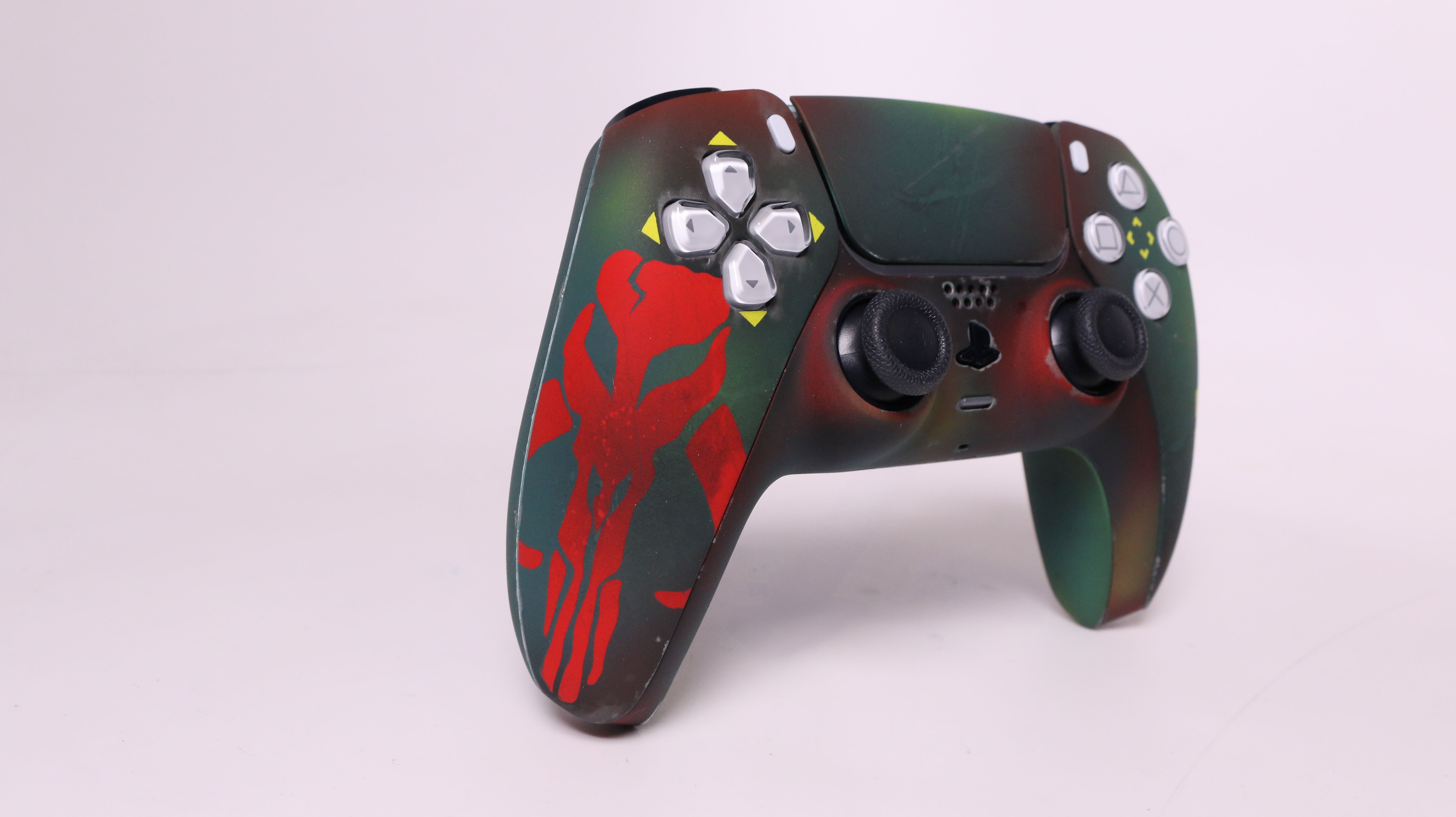A custom-painted PlayStation 5 controller featuring a dark green and black camouflage pattern with a red skull graphic on the left grip and white button accents.