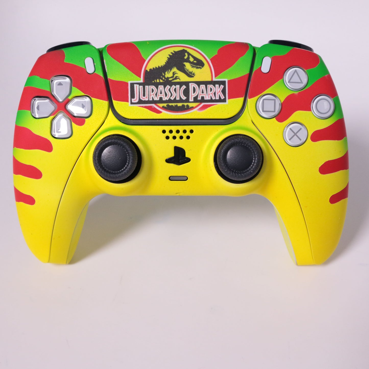 custom-airbrush-painted-dinosaur-theme-ps5-controller-Dino DNA-jeep-Sam Neil-red-yellow-green-TRex A custom PlayStation-style game controller featuring a theme, with a bright yellow body, red and green camouflage patterns, and the  logo on the top center.